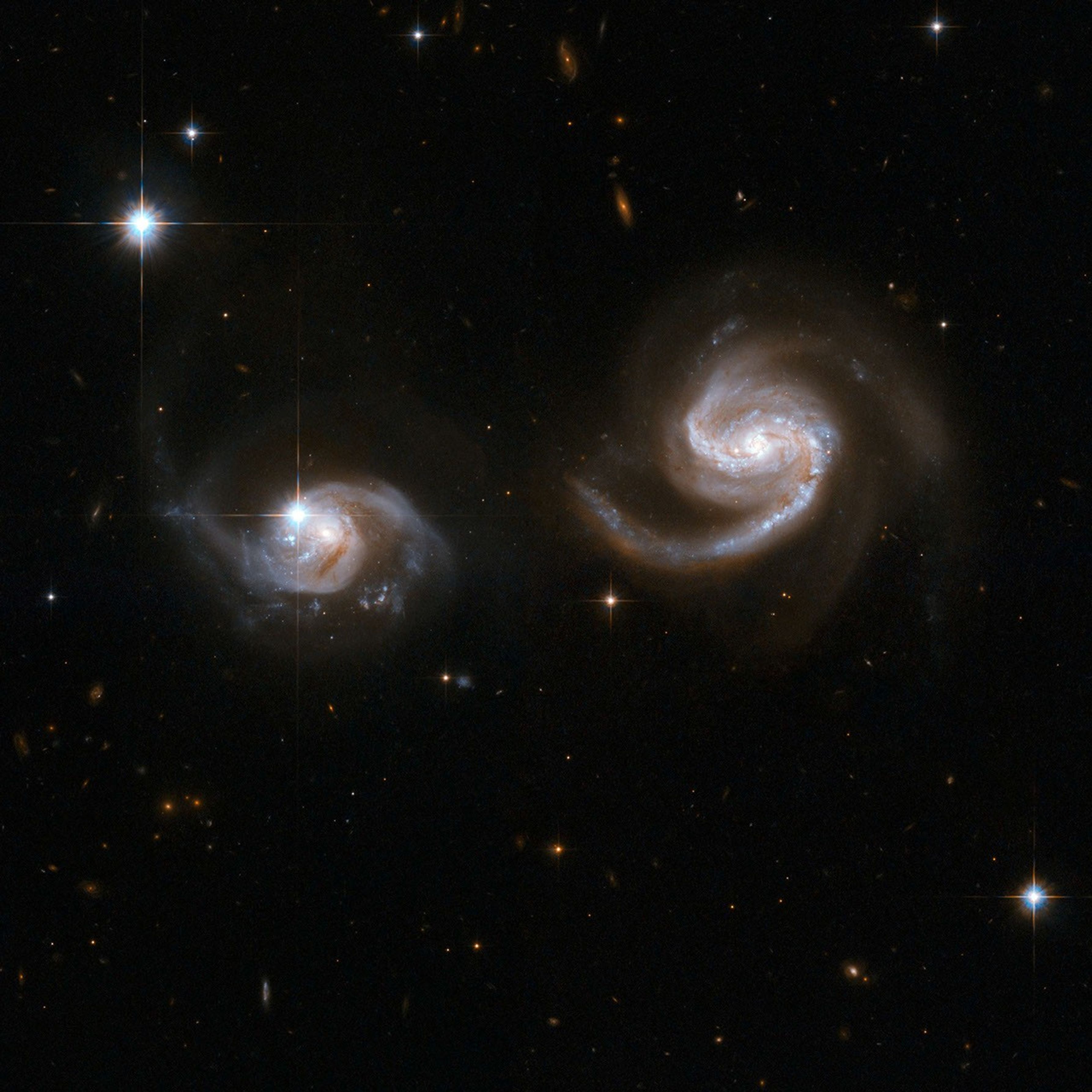 Two spiral galaxies of similar sizes (the one on the right is slightly bigger), looking like a pair of eyes. The right one is white and reddish-orange, the one on the left is mostly white with brown streaks. Foreground stars in the upper left and lower right.